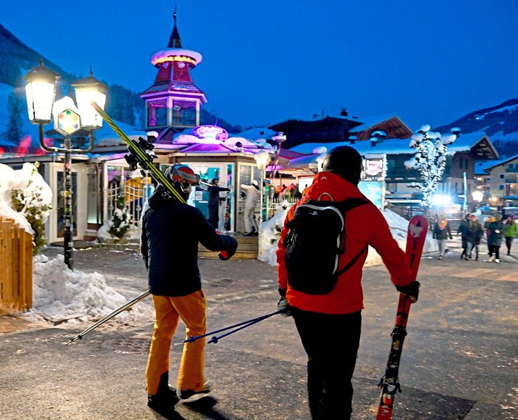 Zwei Touristen auf dem Weg ins Hotel, die Ski auf der Schulter tragend bzw. in der Hand haltend.