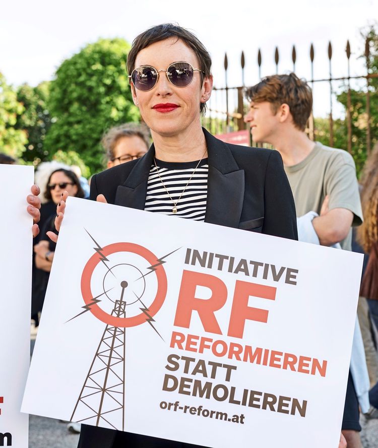Eine Person hält ein Schild mit der Aufschrift „INITIATIVE ORF REFORMIEREN STATT DEMOLIEREN orf-reform.at“. Im Hintergrund sind weitere Personen und ein Zaun zu sehen. Die Szene findet im Freien statt, bei Tageslicht.