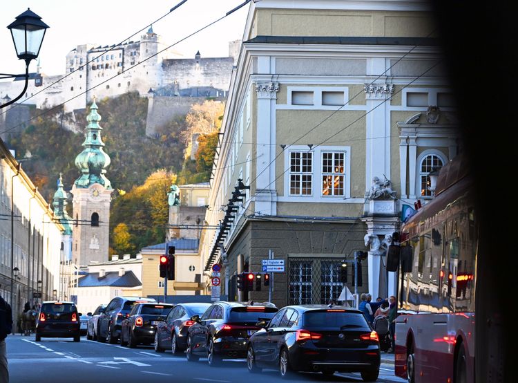 Bauarbeiten am Neutor in Salzburg vor einer steilen Felswand des Mönchsbergs. Ein großer gelber Baukran ragt in die Höhe. Im Vordergrund sind Gebäude, Verkehrsschilder, Autos und Fußgänger zu sehen. Das Bild zeigt die Sanierungs- und Erweiterungsarbeiten für das Festspielzentrum und den Festspielbezirk.