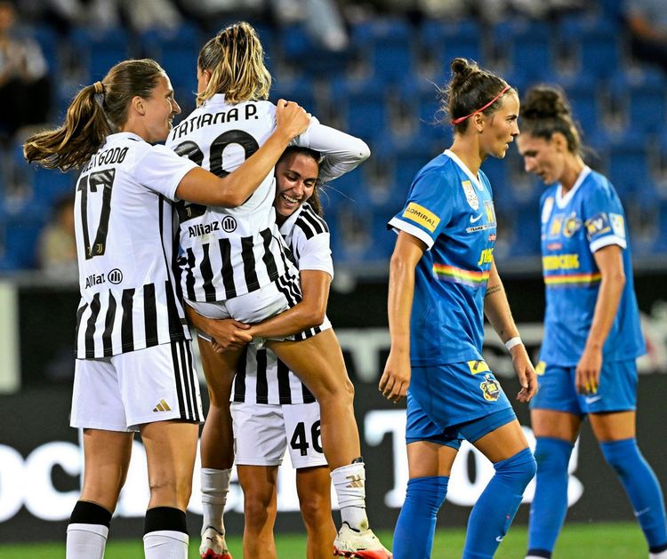 Spielszene aus einem Fußballspiel: Spielerinnen von Juventus Turin in gestreiften Trikots feiern ein Tor; eine Spielerin wird umarmt und hochgehoben. Im Hintergrund laufen Spielerinnen des Gegners SKN St. Pölten in blauen Trikots davon. Stadion und Zuschauer unscharf im Hintergrund.