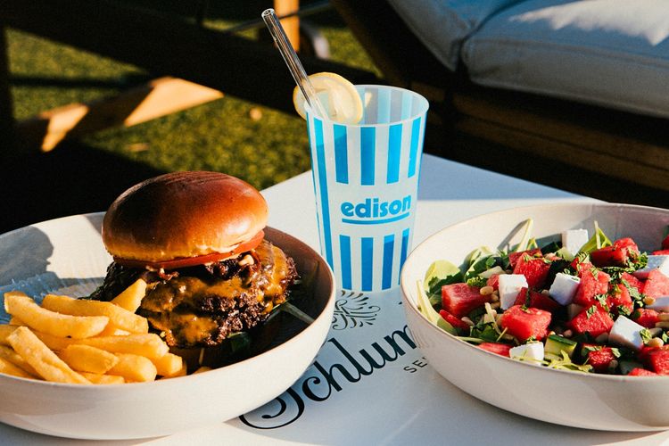 Ein Teller mit einem Cheeseburger und Pommes, daneben eine Schüssel mit einem Salat aus Wassermelone, Feta, Blattgrün und Pinienkernen. Im Hintergrund steht ein gestreifter Becher mit der Aufschrift „edison“ und einer Zitronenscheibe am Strohhalm.