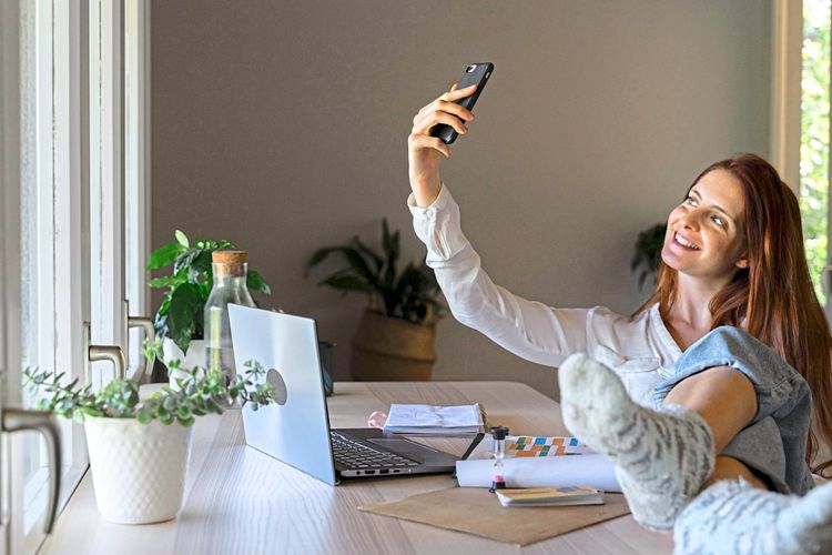 Eine Frau sitzt zu Hause an einem Schreibtisch, lächelt und macht ein Selfie