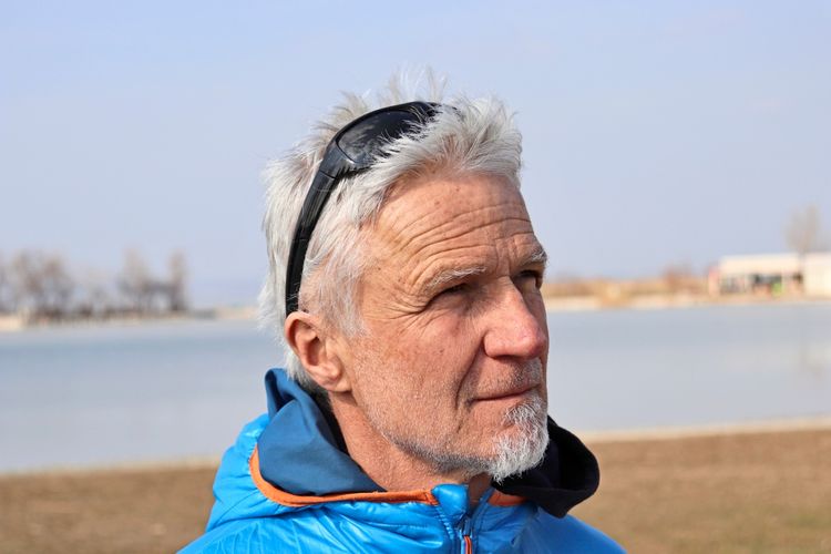 Ein Mann mit weißem Haar trägt eine Sonnenbrille auf dem Kopf und eine blaue Outdoor-Jacke. Der Hintergrund zeigt eine Landschaft mit Wasserfläche und unscharfen Bäumen. Das Gesicht ist unkenntlich gemacht.