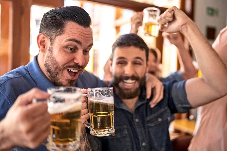 Zwei lachende Männer stoßen in einer fröhlichen Runde mit Bierkrügen in einer Bar an, während sie feiern.