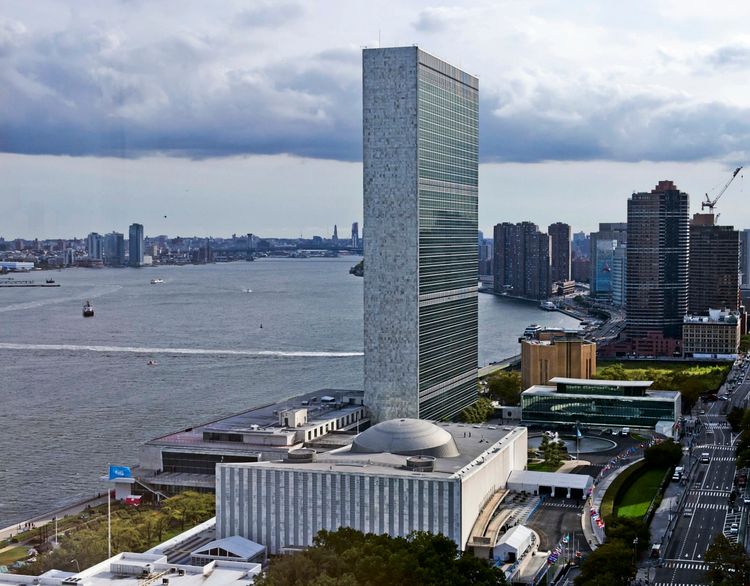 Das Bild zeigt das Hauptquartier der Vereinten Nationen in New York City. Der Turm erhebt sich neben dem East River, umgeben von weiteren Gebäuden und einer Uferpromenade mit wehender UN-Flagge. Im Hintergrund sind weitere Hochhäuser und die Wasserlandschaft zu sehen.