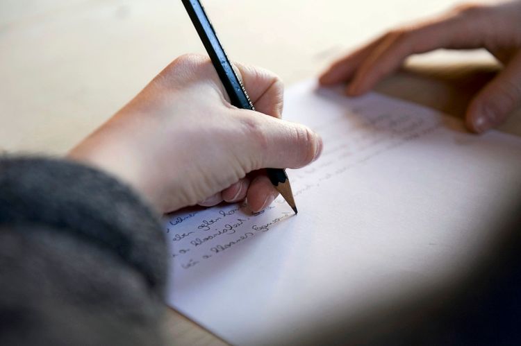 Ein Kind schreibt mit einem Bleistift, den es in der linken Hand hält, auf einem weißen Blatt