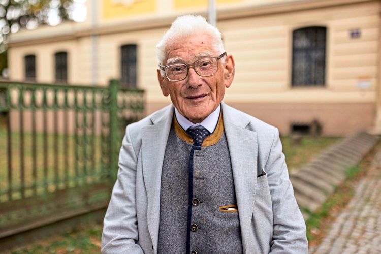Ein älterer Mann mit grauem Anzug, Brille und Krawatte sitzt lächelnd im Freien vor einem Gebäude mit beigem Putz und Metallzaun.