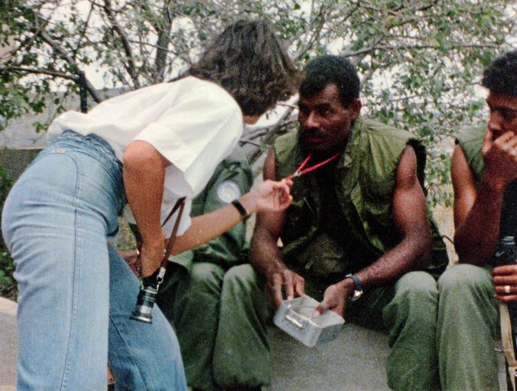 Eine Frau mit einer Kamera um den Hals spricht mit einem Mann in militärischer Kleidung, der auf einer Bank sitzt und eine kleine Box in der Hand hält. Im Hintergrund sind Bäume und weitere Personen zu sehen.