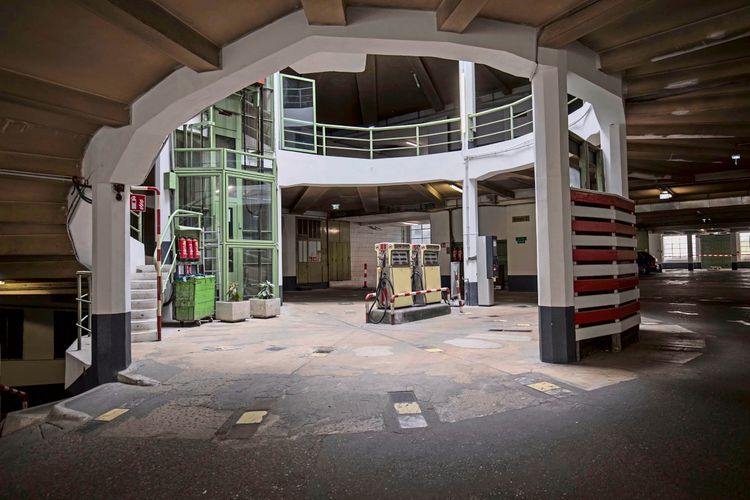 Innenansicht eines mehrstöckigen Parkhauses mit einer zentralen Treppenkonstruktion und einem grünen Aufzug. In der Mitte befinden sich alte, gelbe Zapfsäulen für Kraftstoff. Die Umgebung wirkt industriell mit Betonböden und weiß gestrichenen Säulen.