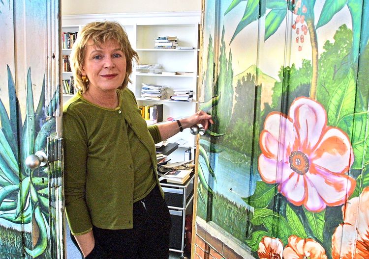 Eine Frau steht in einer geöffneten Tür, die mit buntem, floralen und landschaftlichem Motiv bemalt ist. Im Hintergrund ist ein Büro mit Regalen und Büchern zu sehen. Sie trägt einen grünen Cardigan und blickt freundlich in die Kamera.