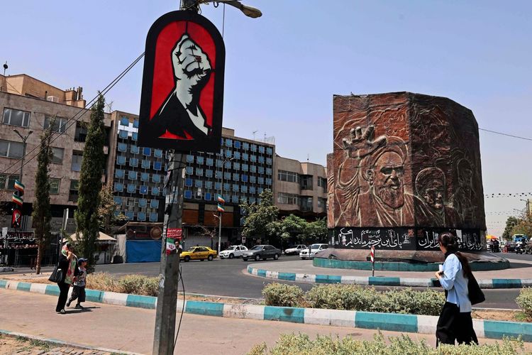 Passanten gehen entlang des Platzes der Islamischen Revolution in Teheran am 21. Juli 2026. Im Vordergrund ist ein Plakat mit einer stilisierten Faust sowie ein monumentales Relief mit Porträts im Hintergrund zu sehen. Gebäude und Fahrzeuge sind ebenfalls sichtbar.