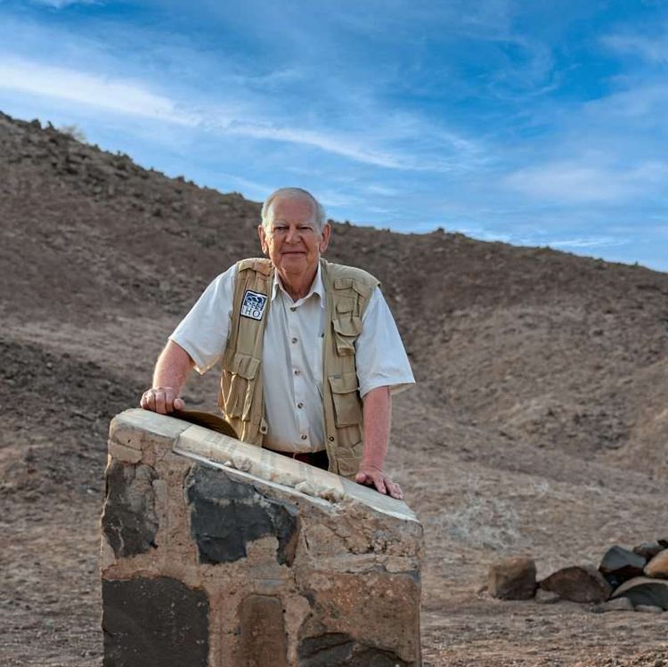 Johanson, ein alter Mann mit weißen Haaren, stützt sich in der kargen Landschaft auf einen Gedenkstein.