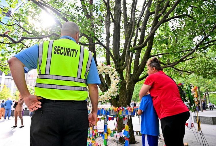 Ein Sicherheitsmitarbeiter mit gelber Weste steht vor einem Baum, dem sogenannten „Survivor Tree“, der die Anschläge vom 11. September überlebt hat. Links im Hintergrund sind weitere Personen zu sehen. Eine Frau in rotem Shirt umarmt ein Kind vor dem Baum, der mit Blumen und bunten Bändern geschmückt ist.