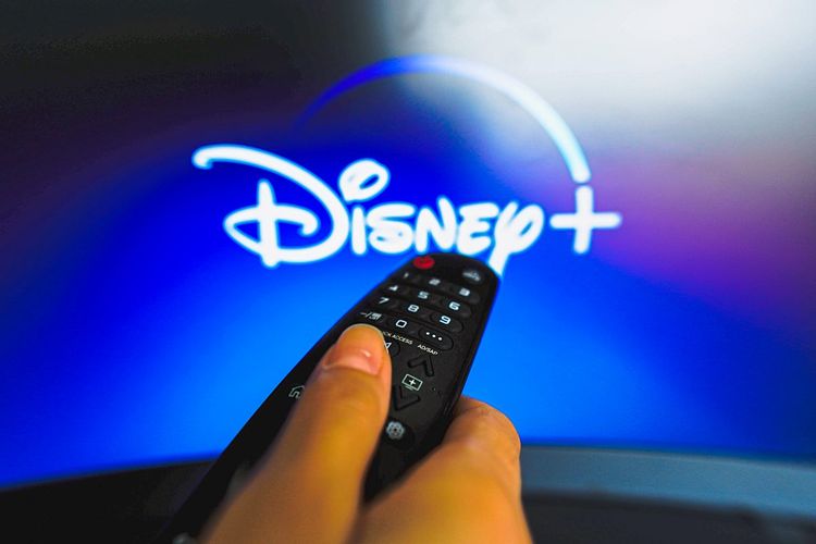 Auf diesem Foto ist eine Nahaufnahme einer Hand zu sehen, die eine TV-Fernbedienung vor dem Disney-Plus-Logo hält.