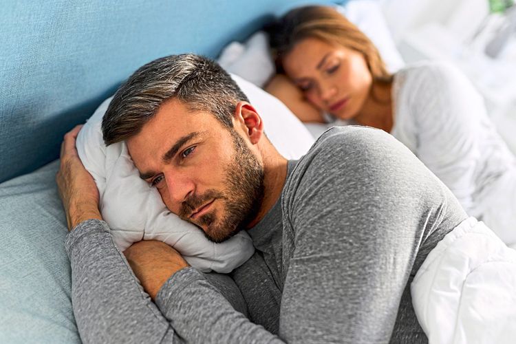Ein besorgt wirkender Mann liegt mit nachdenklichem Gesicht im Bett, mit dem Rücken zu einer im Hintergrund schlafenden Frau