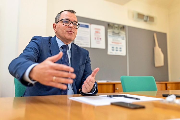 Ein Mann in einem Anzug sitzt an einem Tisch und gestikuliert mit den Händen, als ob er etwas erklärt. Auf dem Tisch liegen Papiere und ein Mobiltelefon. Im Hintergrund sind eine Pinnwand mit Plakaten und eine Stofftasche zu sehen.