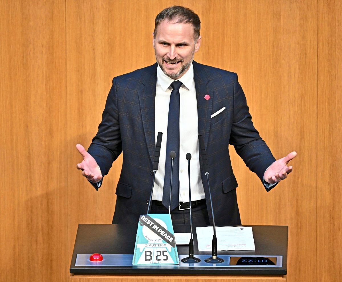 Ein Mann in einem dunklen Anzug mit Krawatte steht an einem Rednerpult während einer Sitzung im österreichischen Parlament in Wien. Vor ihm befinden sich Mikrofone, ein roter Knopf und ein Schild mit einer Vignette und der Aufschrift 