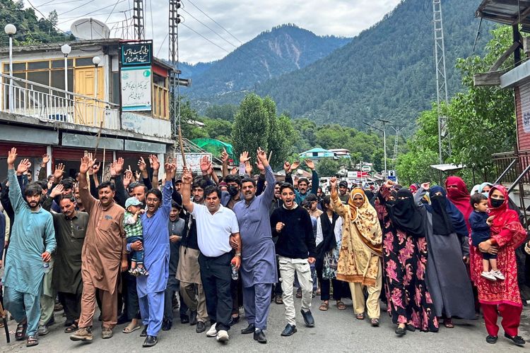 Menschen protestieren in einer Gruppe auf einer Straße im Neelum Valley, Pakistanisch-Kaschmir. Sie heben die Hände und rufen slogans. Im Hintergrund sind Gebäude, grüne Bäume und bewaldete Berge zu sehen.