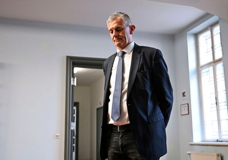 Ein Mann in einem Anzug mit Krawatte steht in einem hellen Raum vor einer Tür. Im Hintergrund sind ein Fenster und Schilder an der Wand zu sehen.