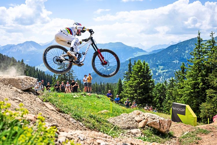 Ein Mountainbiker, gekleidet in weißer Ausrüstung, springt während eines Downhill-Rennens über eine steinige Strecke. Im Hintergrund stehen Zuschauer, und die Szene ist von einer malerischen Berglandschaft umgeben. Ein Schild mit der Aufschrift 