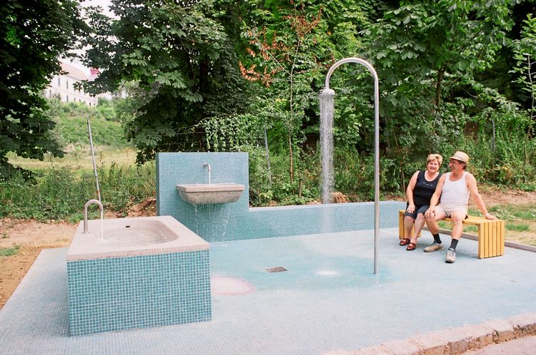 Ein öffentlicher Badebereich in Loosdorf mit einer Duschsäule, einem Becken und einer Wasserstelle, gestaltet mit blauen Fliesen. Zwei Personen sitzen lächelnd auf einer Holzbank am Rand. Im Hintergrund sind Bäume und ein Gebäude zu sehen. Kunstwerk von Iris Andraschek.