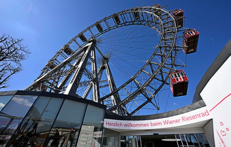Das Bild zeigt das Wiener Riesenrad im Prater in Wien vor einem klaren blauen Himmel. Im Vordergrund ist der Eingangsbereich mit der Aufschrift „Herzlich willkommen beim Wiener Riesenrad“ zu sehen.