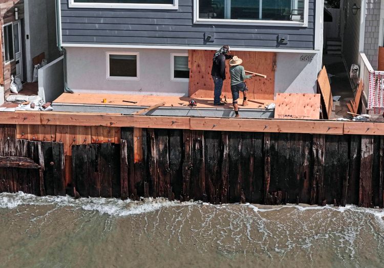 Zwei Personen arbeiten an der Absicherung eines Hauses nahe Belmont Shore Beach in Long Beach, Kalifornien. Sie befestigen Holzplatten, um die Fenster und unteren Bereiche des Gebäudes zu schützen. Im Vordergrund sind eine Ufermauer sowie Wasserwellen zu sehen.