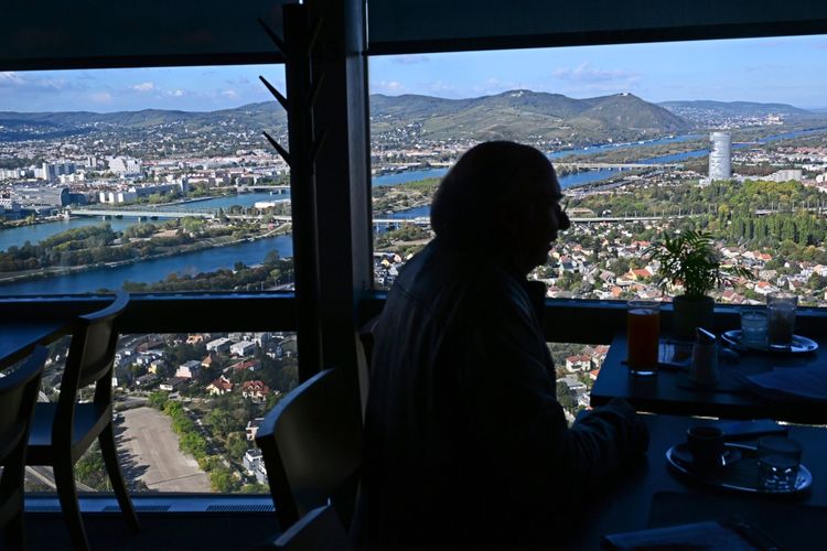 Dimitré Dinev sitzt silhouettenhaft in einem Restaurant mit Panoramablick auf eine Stadtlandschaft. Im Hintergrund fließen mehrere Flussarme durch die Stadt, umgeben von Gebäuden, Hügeln und Grünflächen. Ein markanter, moderner Turm ist rechts im Bild zu sehen.