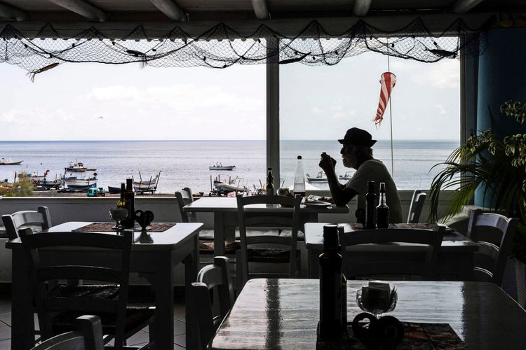 Ein Mann sitzt in einem Restaurant auf der Insel Stromboli, Italien, mit Blick auf das Meer und Boote im Hintergrund. Die Einrichtung des Restaurants ist schlicht, mit weißen Holzmöbeln und Fischernetzen als Dekoration.