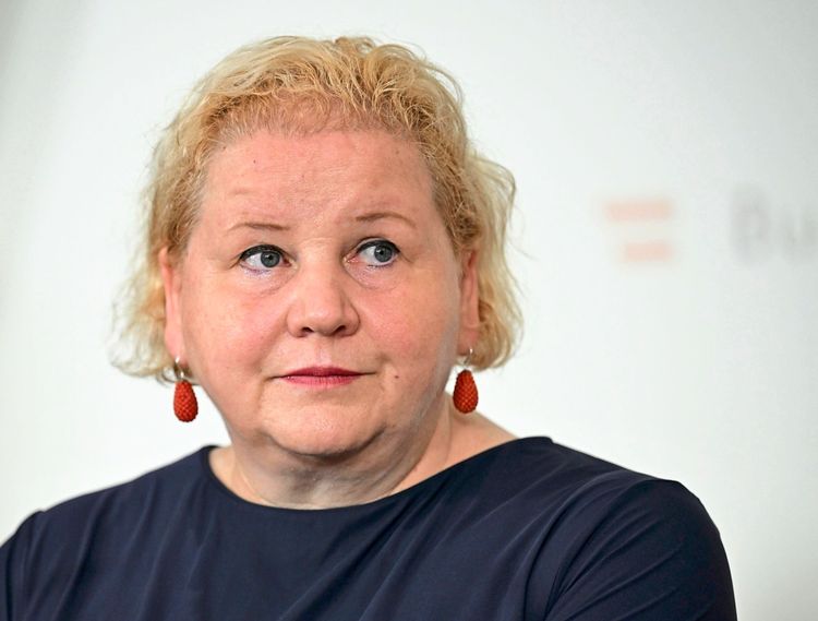 Eine Frau mit blonden Haaren und roten Ohrringen, bekleidet mit einem dunkelblauen Oberteil, blickt nachdenklich zur Seite. Der Hintergrund ist neutral.