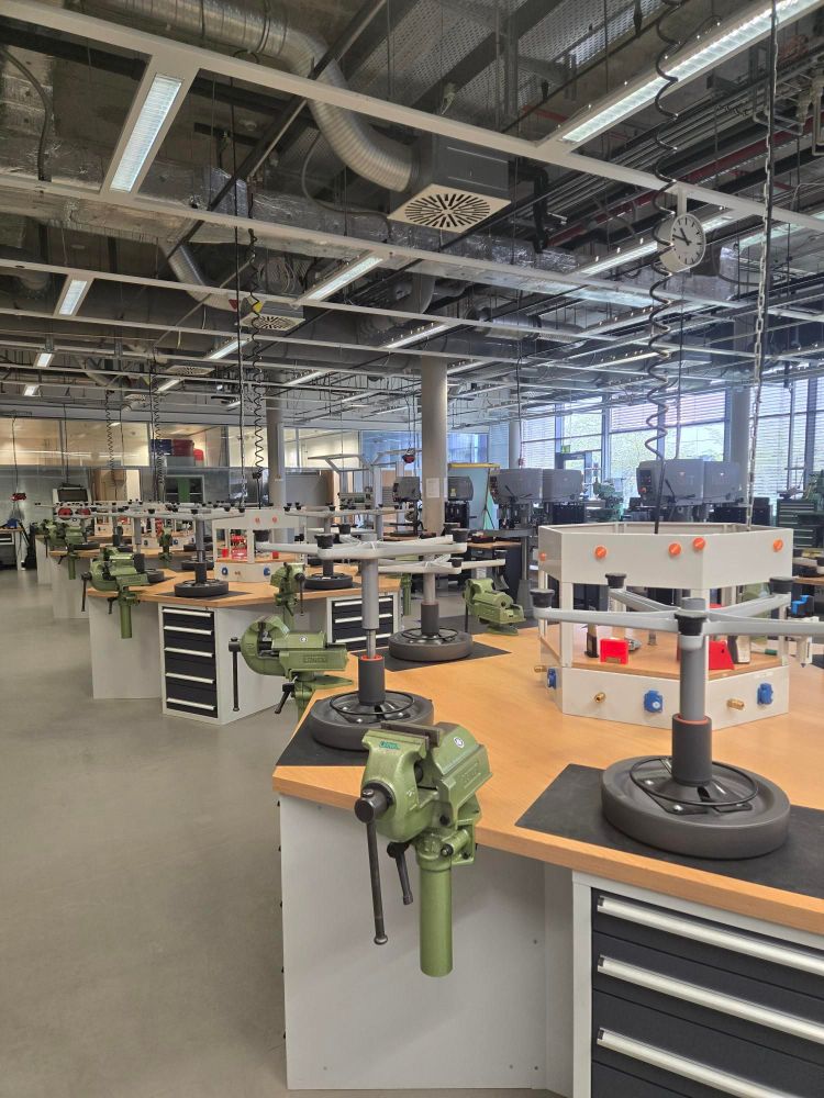 Eine Werkshalle mit Tischen, Schraubstücken, umgedrehten Hockern. An der Decke sieht man Leitungsrohre und Kabel.