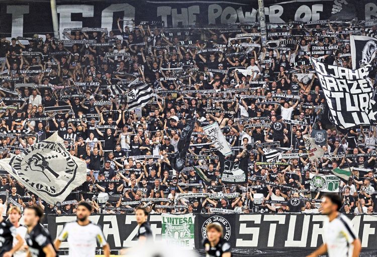 Sturm-Graz-Fans jubeln begeistert mit Schals, Bannern und Fahnen während eines Fußballspiels im Stadion Graz-Liebenau. Im Vordergrund sind einige Spieler auf dem Spielfeld zu sehen.