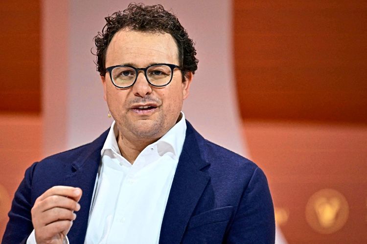 Ein Mann mit lockigem dunklem Haar und Brille spricht, während er auf einer Bühne steht. Er trägt ein weißes Hemd unter einem dunkelblauen Sakko. Der Hintergrund ist unscharf, mit orangefarbenen und weißen Tönen.