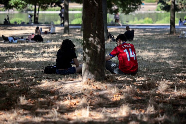 Ein Paar sitzt im Schatten eines Baumes auf ausgedörrtem Gras im Bois de Vincennes Park in Paris während einer Hitzewelle. Der Mann trägt ein rotes Trikot mit der Aufschrift „D. Doué 14“.