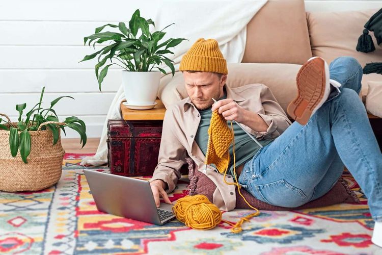 Junger Mann mit Strickzeug sitzt auf dem Boden und schaut im Internet nach, wie es funktioniert