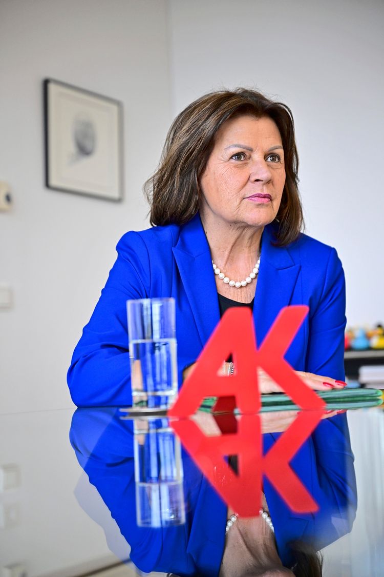 Eine Frau in einem blauen Blazer und mit einer Perlenkette sitzt an einem Tisch. Vor ihr stehen ein Glas Wasser und ein rotes 