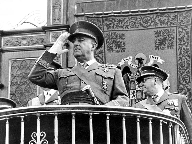 Schwarz-Weiß-Fotografie eines Militärs in Uniform, der auf einem Balkon steht und salutiert. Im Hintergrund ist ein kunstvoll verzierter Wandteppich mit Emblemen und Wappen zu sehen. Weitere uniformierte Personen stehen neben ihm.