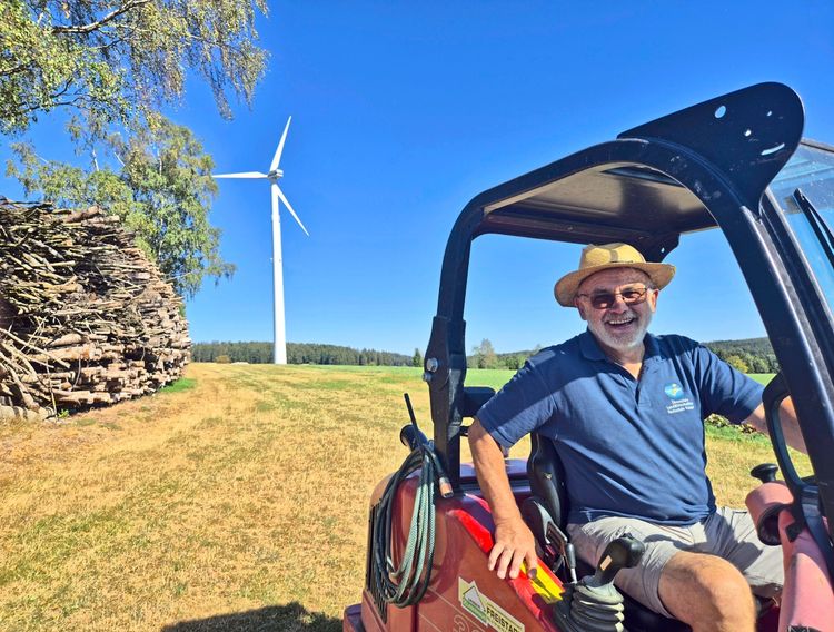 Ein Mann mit Strohhut und dunkelblauem Poloshirt sitzt lächelnd in einem Roten Traktor auf einer Wiese. Im Hintergrund befinden sich ein Windrad, ein Stapel Holz und Bäume unter einem klaren blauen Himmel.