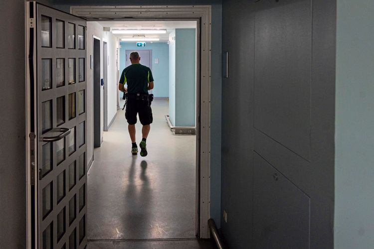 Ein Wachmann in sportlicher Kleidung mit Ausrüstungsgürtel geht durch einen Flur einer Haftanstalt. Im Vordergrund ist eine geöffnete Zellentür zu sehen, der Flur ist hell erleuchtet.