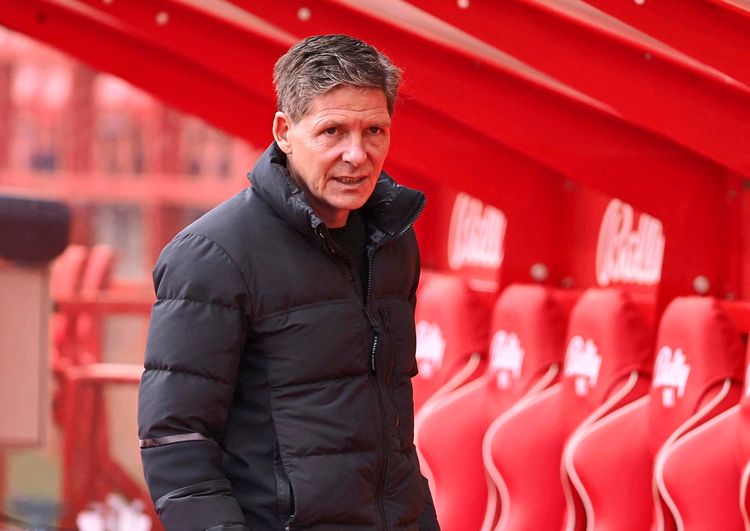 Ein Mann in einer schwarzen Daunenjacke steht am Spielfeldrand vor roten Sitzbänken. Im Hintergrund sind unscharf rote Strukturen zu sehen.
