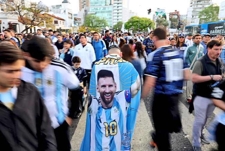Eine Menschenmenge von Fußballfans vor einem Stadion. Ein Fan trägt eine argentinische Flagge mit einem Bild von Lionel Messi und der Nummer 10 darauf. Hintergrund mit Gebäuden und Bäumen.