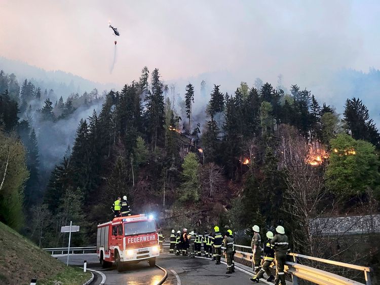 Ein Waldbrand im Lesachtal, Österreich: Im Vordergrund stehen Feuerwehrleute neben einem Feuerwehrfahrzeug, während Löscharbeiten durchgeführt werden. Im Hintergrund brennt der Wald, Flammen sind sichtbar. Am Himmel fliegt ein Helikopter und lässt Wasser aus einem Löschbehälter ab. Der Himmel ist rauchverhangen.