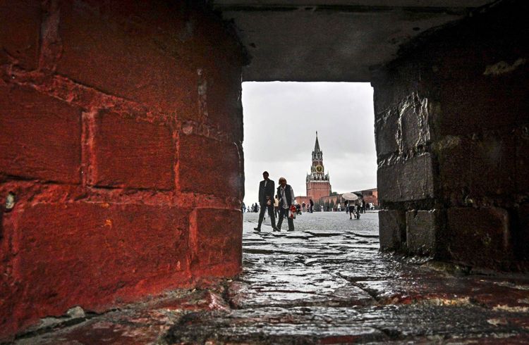 Menschen gehen vor der Spasskaja-Turm auf dem Roten Platz in Moskau entlang. Der Blick erfolgt durch eine schmale Öffnung in einer roten Ziegelmauer. Es ist ein bewölkter Tag.