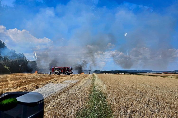 Ein Feldbrand in einer ländlichen Region, bei dem dichter Rauch und Flammen sichtbar sind. Ein Feuerwehrauto und Feuerwehrleute arbeiten an den Löscharbeiten. Im Hintergrund sind blauer Himmel, Wolken und angrenzende Felder zu sehen.