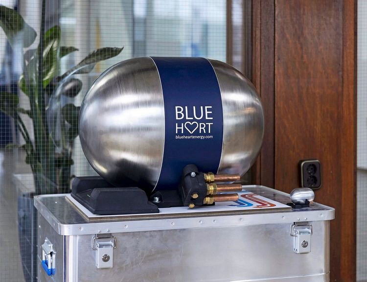 Metallisches Gerät mit ovaler Form und der Aufschrift „BLUE HEART“ sowie „blueheartenergy.com“. Das Gerät ist auf einer silbernen Kiste montiert, mit sichtbaren Kupferrohren und schwarzer Halterung. Im Hintergrund sind eine Glastür, eine Topfpflanze und Holzelemente zu sehen.
