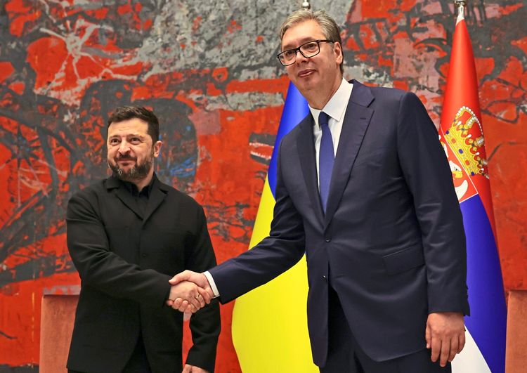 Zwei Männer stehen vor einem roten Hintergrund mit abstraktem Muster und schütteln sich die Hand. Im Hintergrund sind die Flaggen der Ukraine und Serbiens sichtbar.