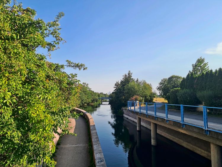 Ein idyllischer Fluss, flankiert von dichtem grünen Gebüsch und Bäumen. Rechts führt ein Geh- und Radweg mit blauen Geländern auf Stelzen am Wasser entlang. Links verläuft ein schmaler Pfad. Der Himmel ist klar und blau.