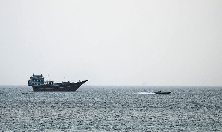 Ein großes Schiff liegt ruhig im Wasser der Straße von Hormus, während ein kleines Motorboot vorbeifährt. Der Himmel ist dunstig.