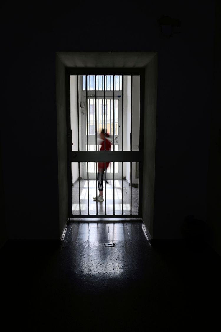 Eine geschlossene Zellentür mit Stahlgittern in einem Gefängnis. Dahinter ist unscharf eine Person in einem roten Oberteil zu erkennen, die in einem erleuchteten Gang steht. Die Umgebung ist dunkel, nur der Bereich hinter der Tür ist beleuchtet.