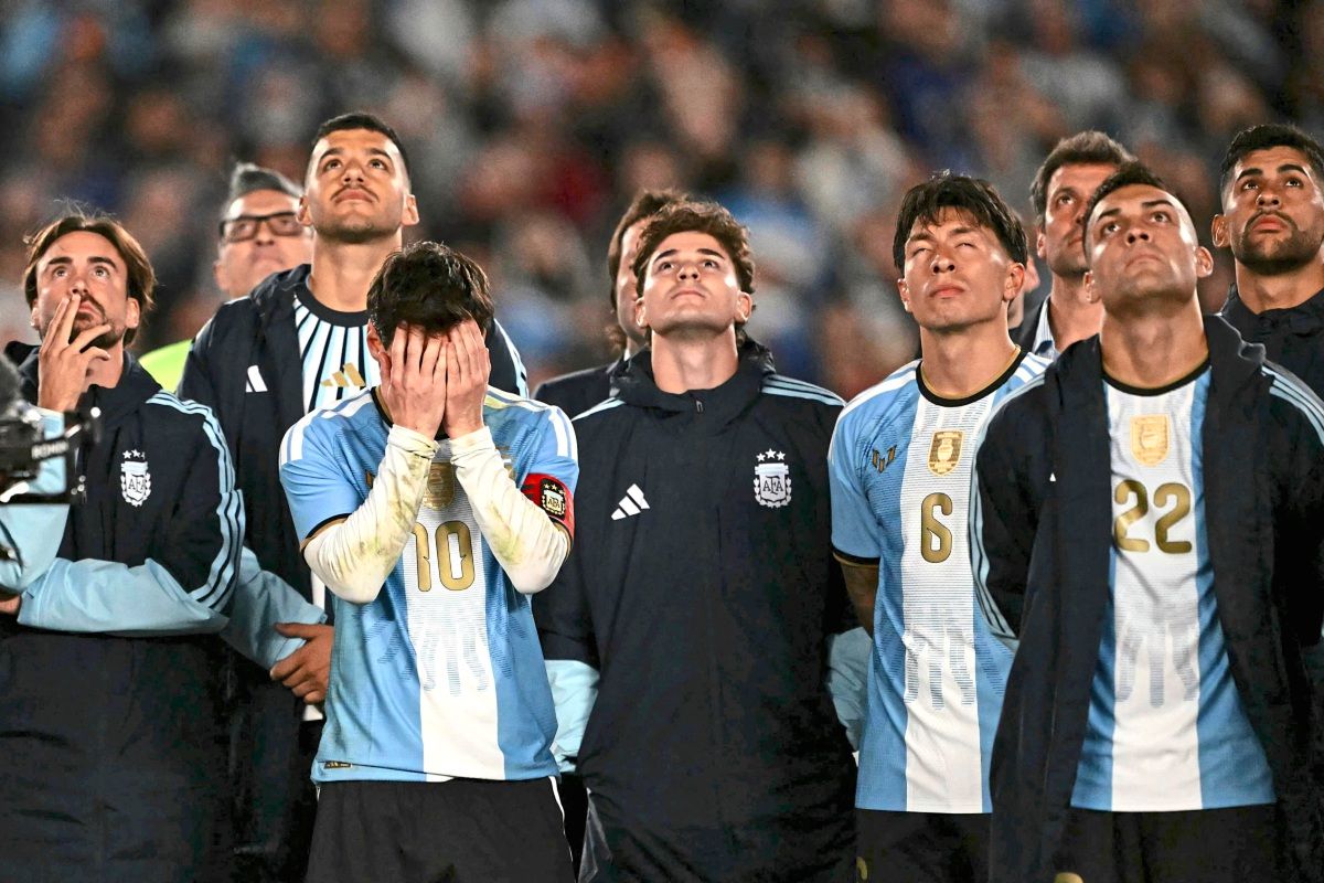 Argentinische Fußballspieler, darunter ein Spieler mit der Nummer 10, stehen in Nationaltrikots aufgereiht. Der Spieler mit der Nummer 10 bedeckt sein Gesicht mit den Händen, während andere Spieler nachdenklich oder emotional nach oben schauen.